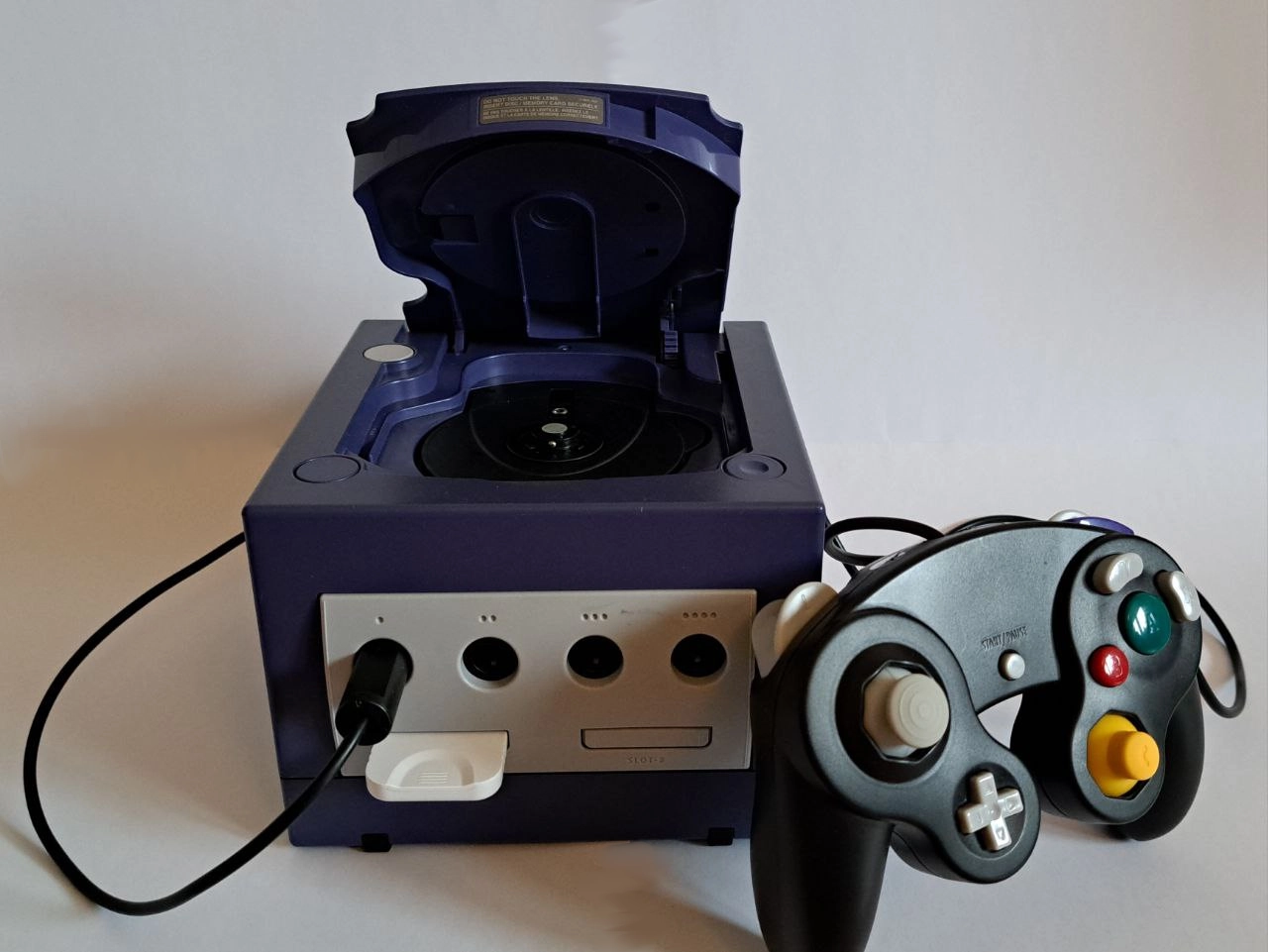 Lila Gamecube auf weißem Hintergrund. Angeschlossen sind eine weiße Memorycard in Slot 1 sowie ein schwarzer Controller in Anschluss 1. Die Klappe des Gamecubes ist offen.