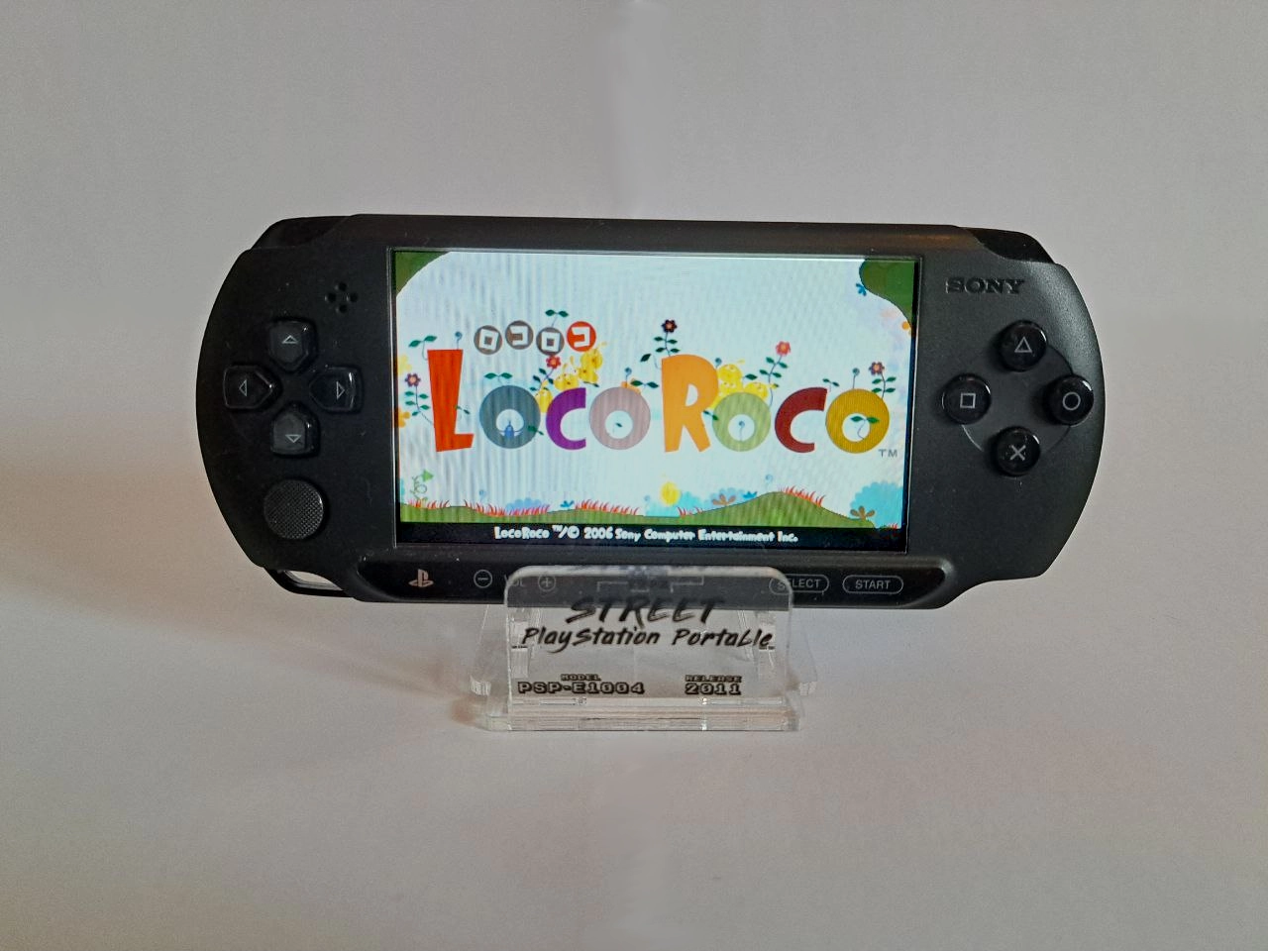 Eine schwarze PSP Street auf weißem Hintergrund. Die PSP ist eingeschaltet und zeigt den Startbildschirm des Spiels Loco Roco.