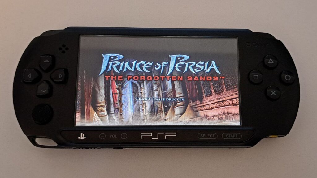 Eingeschaltete PSP auf weißem Untergrund. Zu sehen ist der Startbildschirm von Prince of Persia: The forgotten Sands