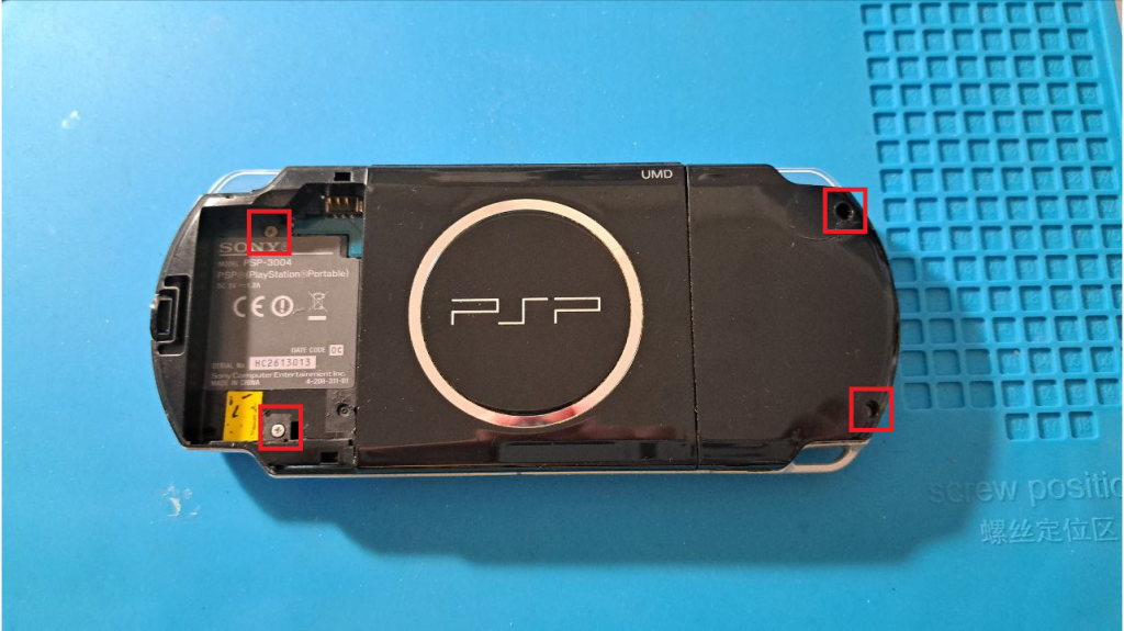 Schwarze PSP 3000 auf blauer Lötmatte. Akkuklappe und Akku sind entfernt. Vier Schrauben sind rot markiert.